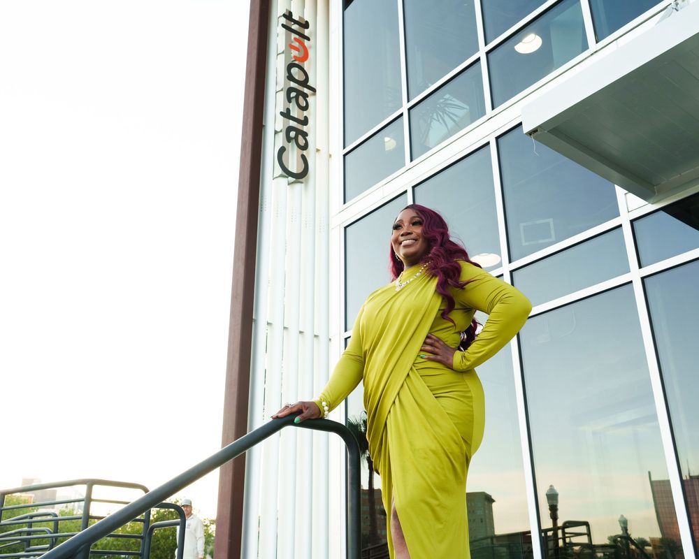 Woman in yellow-green dress poses confidently outside Catapult building, featuring glass facade and modern architecture.