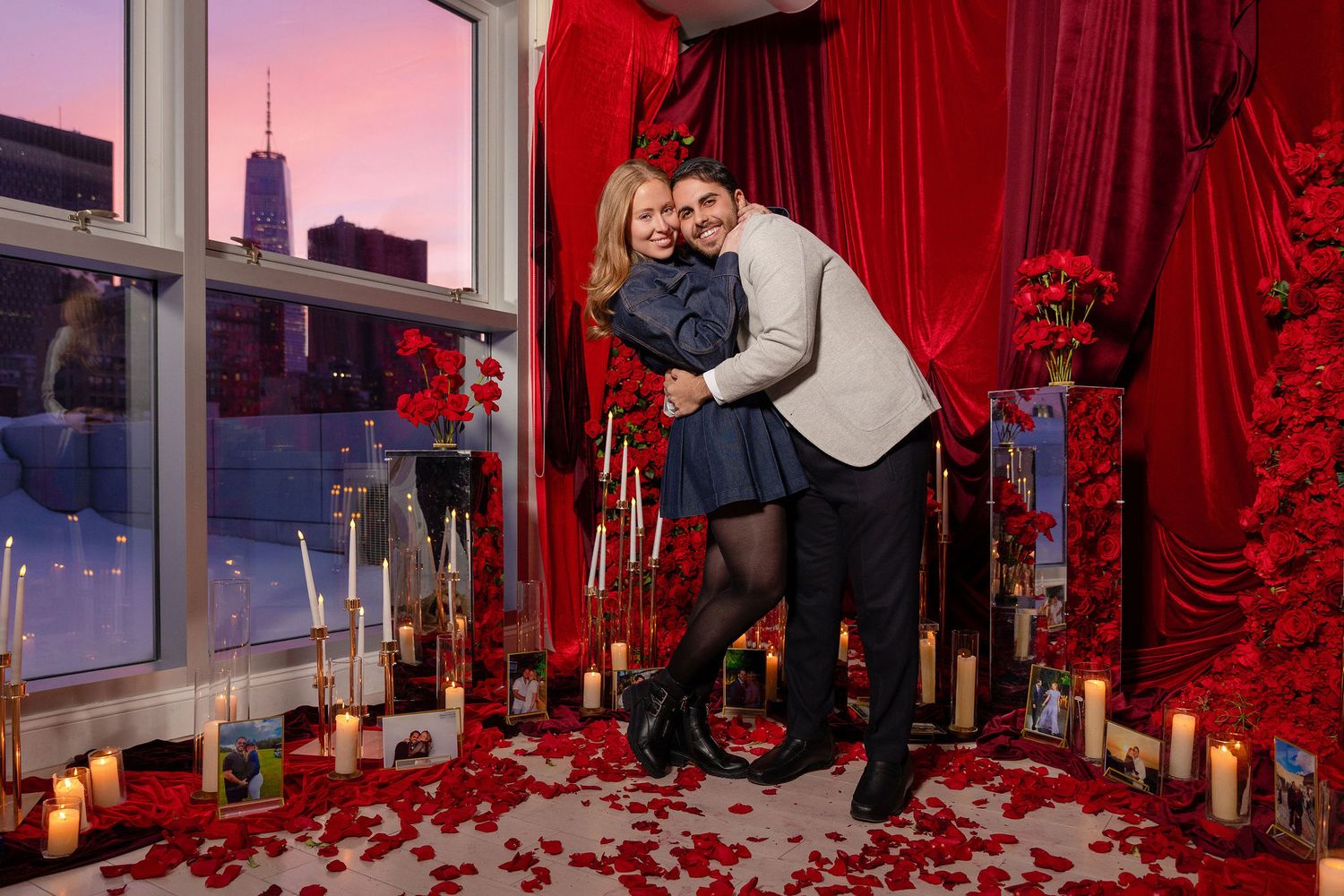 Couple embracing in romantic NYC room with red rose petals, candles, and red drapes at sunset.