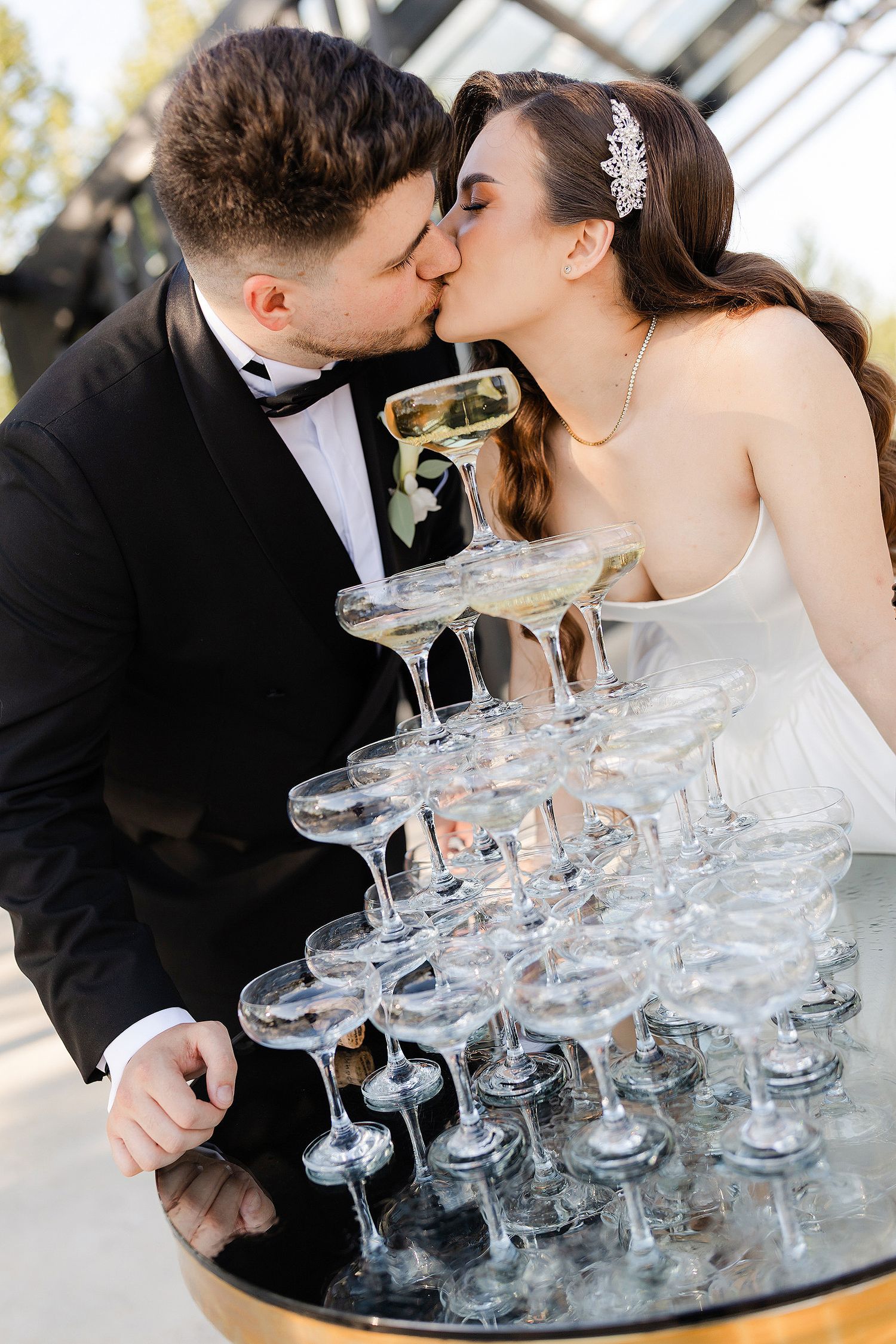 Bride and groom kissing while pouring champagne into a elegant tower of coupe glasses at their wedding reception.