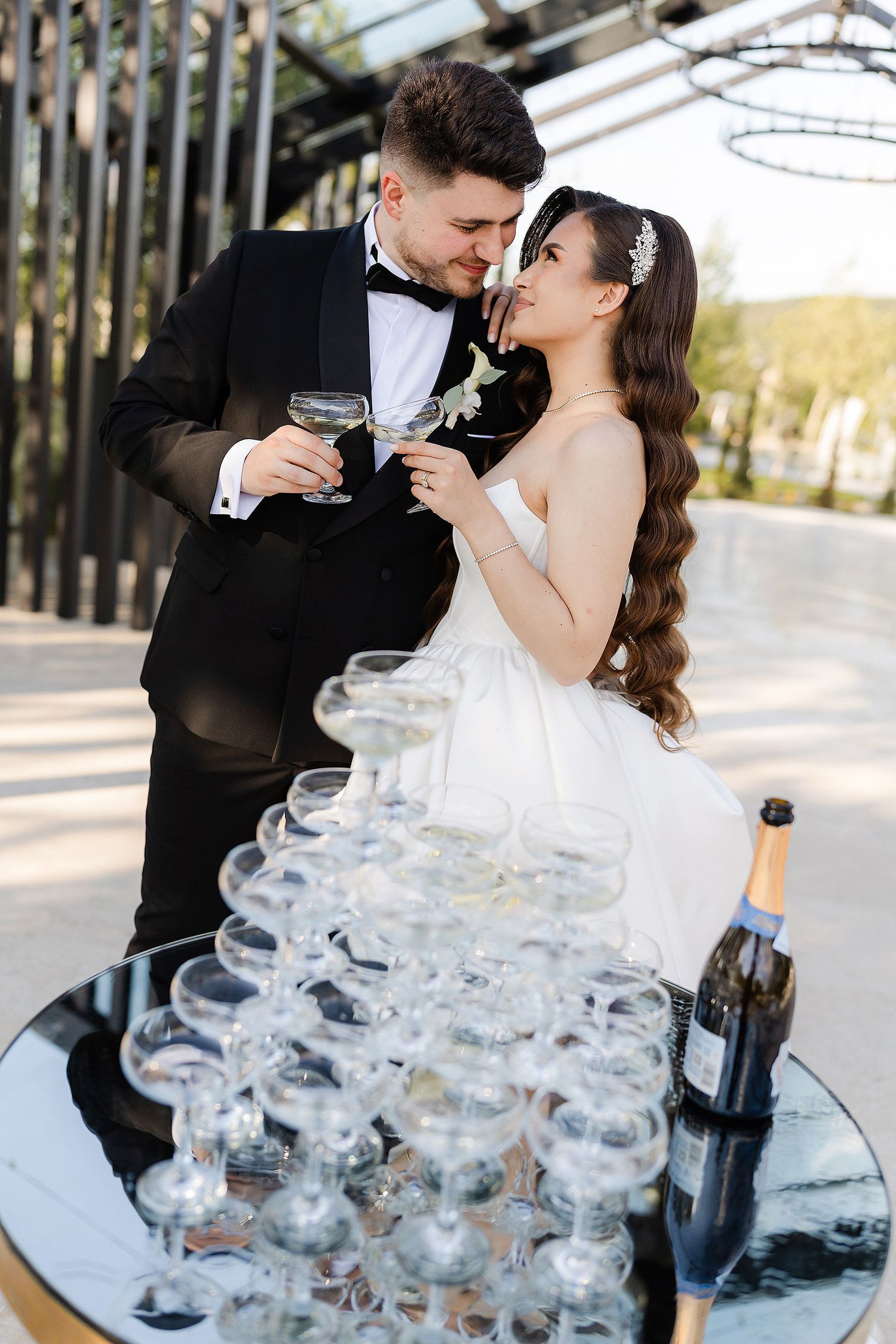 Bride and groom toasting champagne glasses beside a pyramid of glasses at their elegant outdoor wedding celebration.