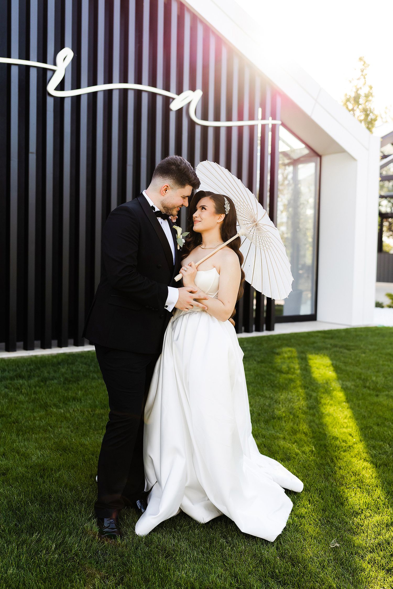 Bride and groom embrace on green lawn, she holds white parasol, he wears black tuxedo, modern building backdrop.