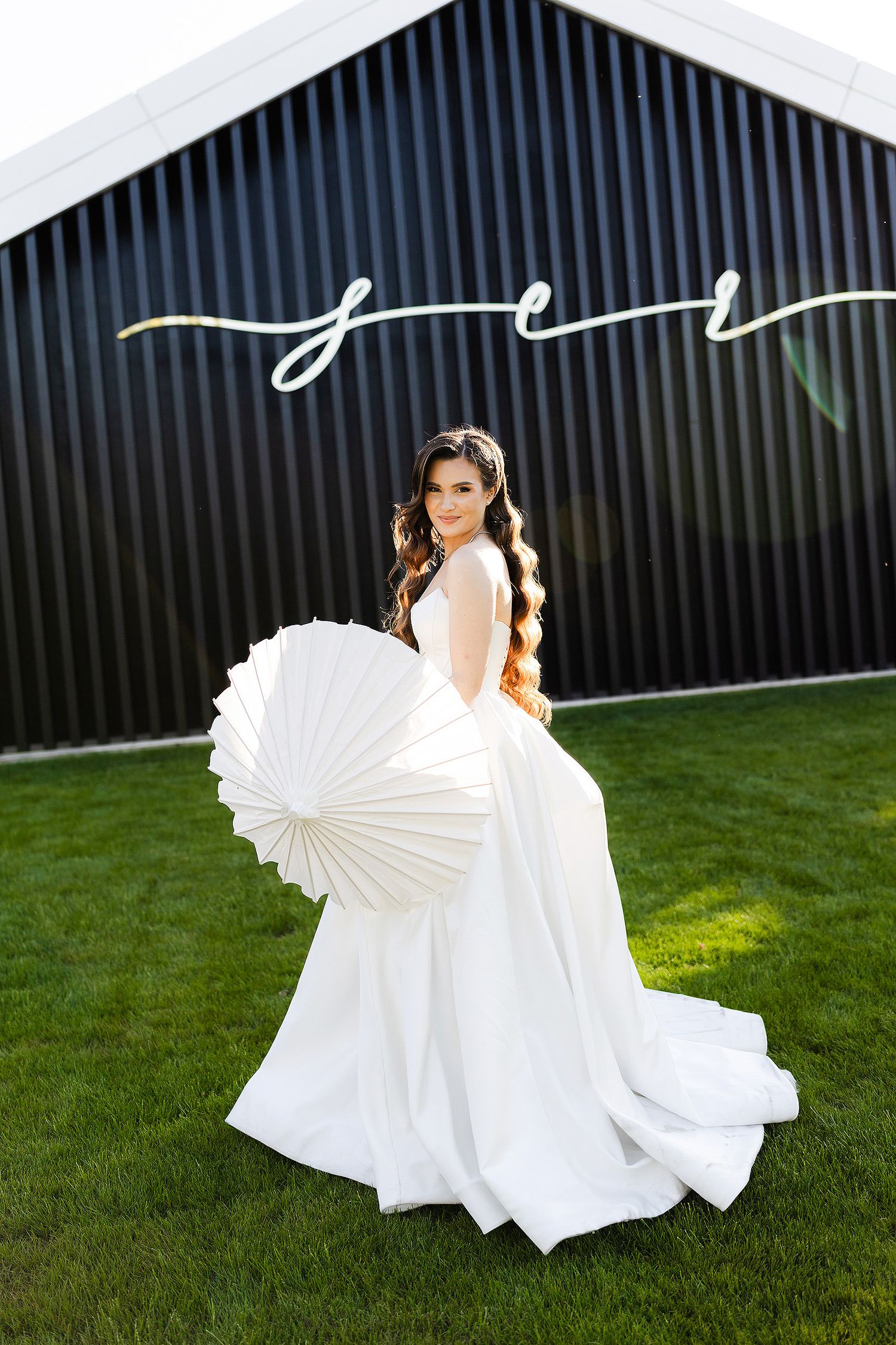Bride in white gown holding paper parasol on green lawn beside black barn with cursive neon sign.