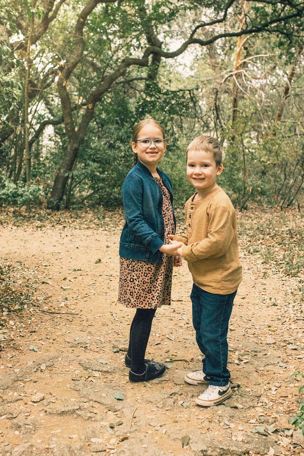 Two people stand holding hands on a dirt path surrounded by trees and autumn foliage in a wooded area.