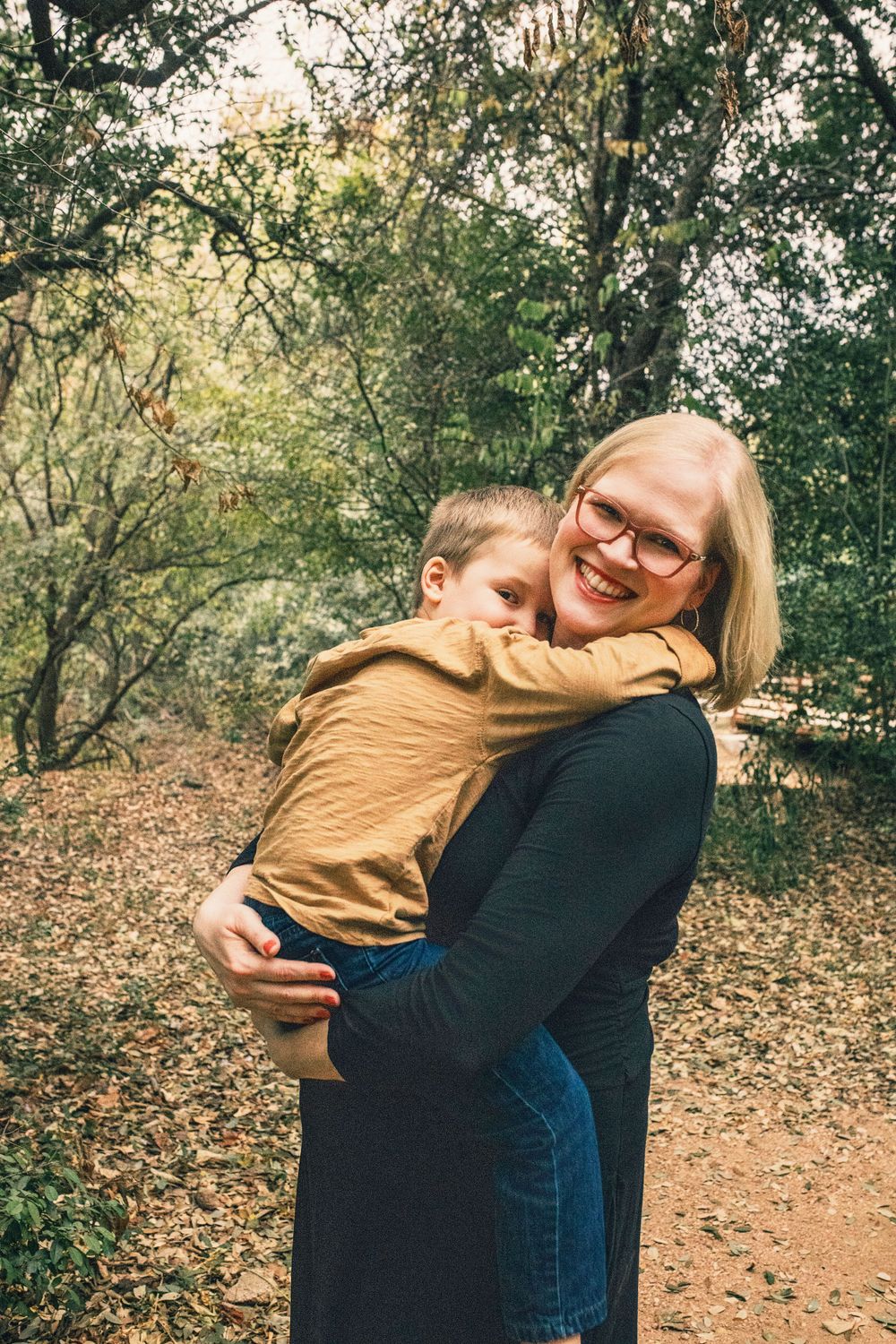 Two figures embrace while standing in a forest clearing with autumn leaves on the ground.