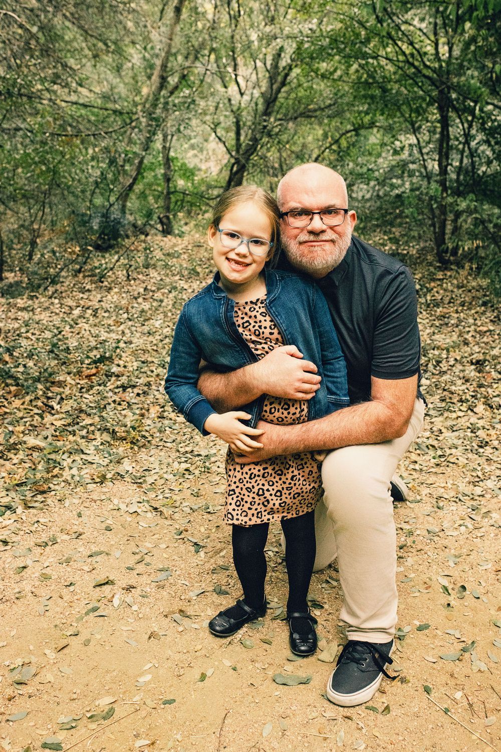 An adult and child pose together on a dirt trail in the woods wearing casual attire and glasses.