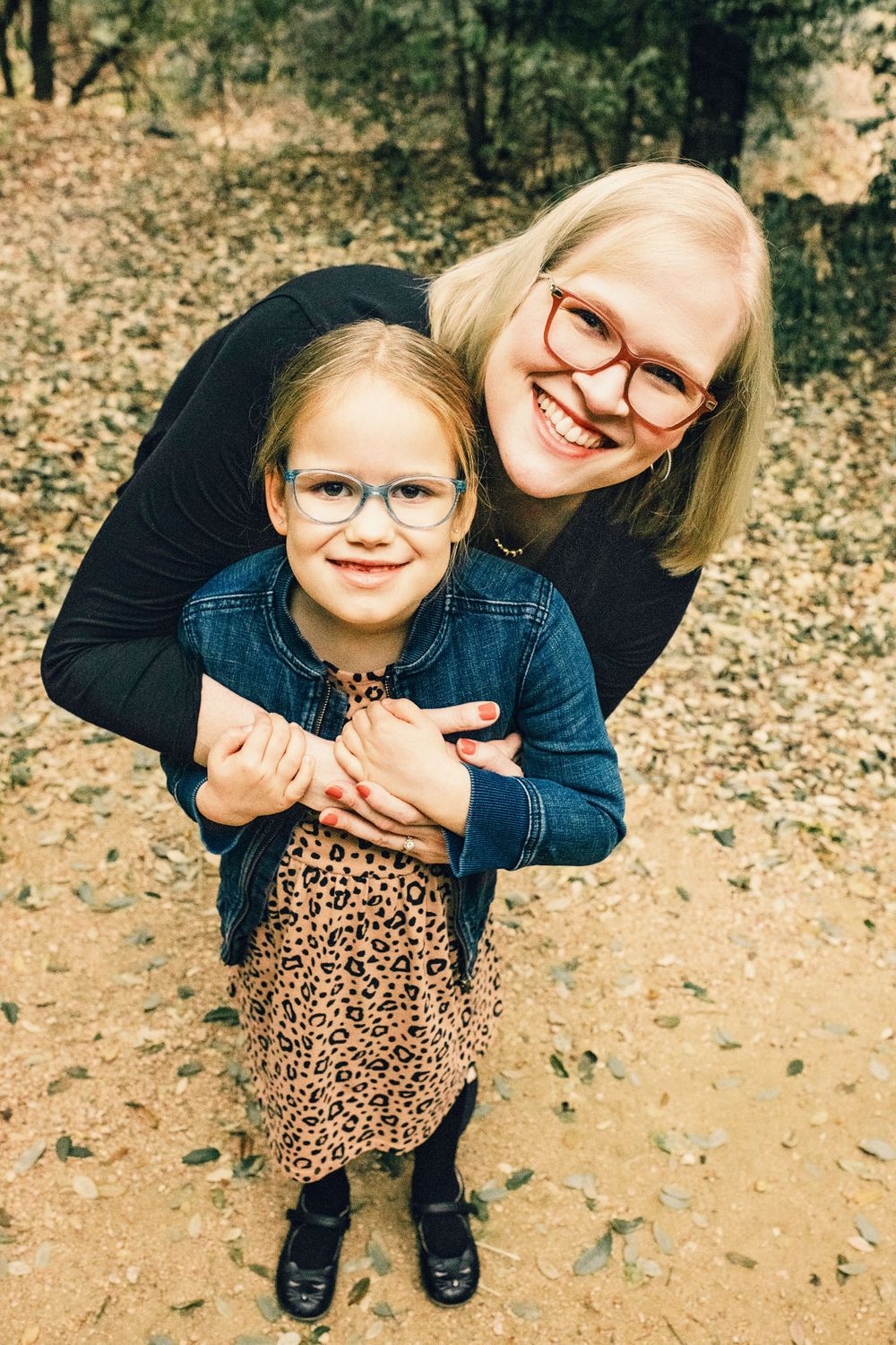 A mother and daughter wearing matching glasses embrace while smiling outdoors on a fall day with leaves on the ground.