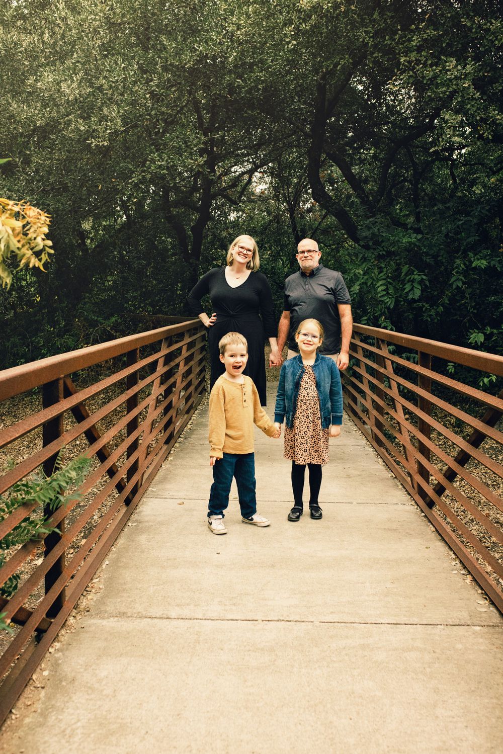 A family stands together on a wooden footbridge surrounded by lush green trees in a natural setting.