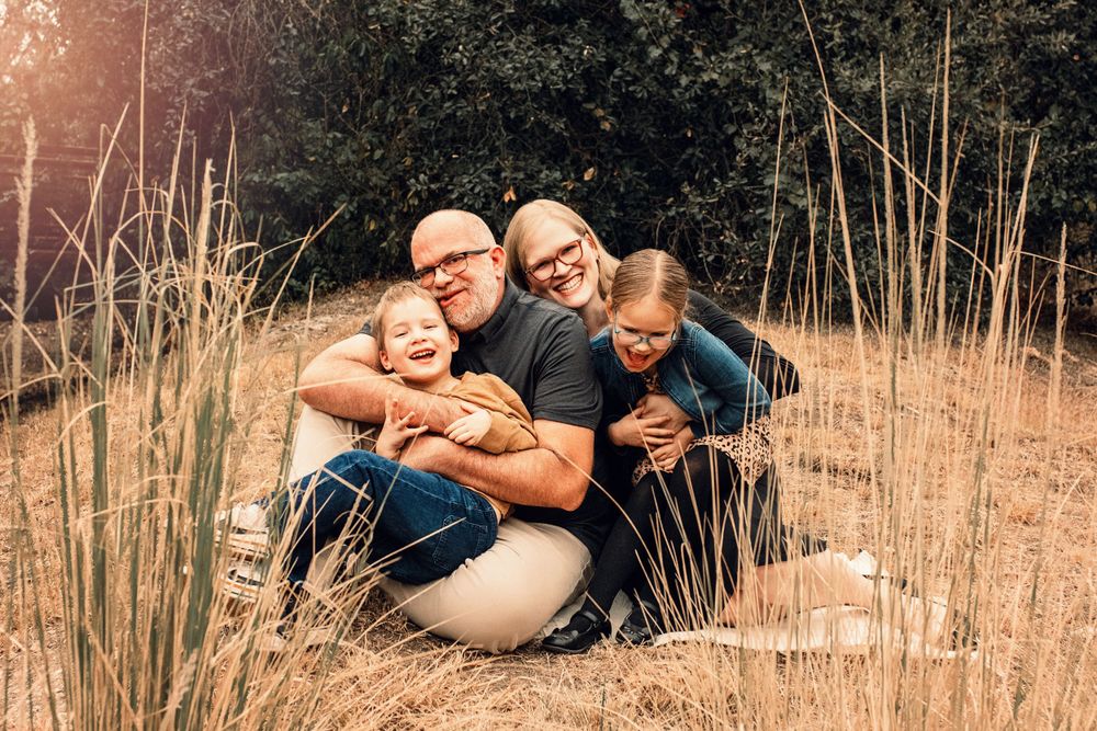 A loving family embraces while sitting in tall grass during a warm sunset photo session.
