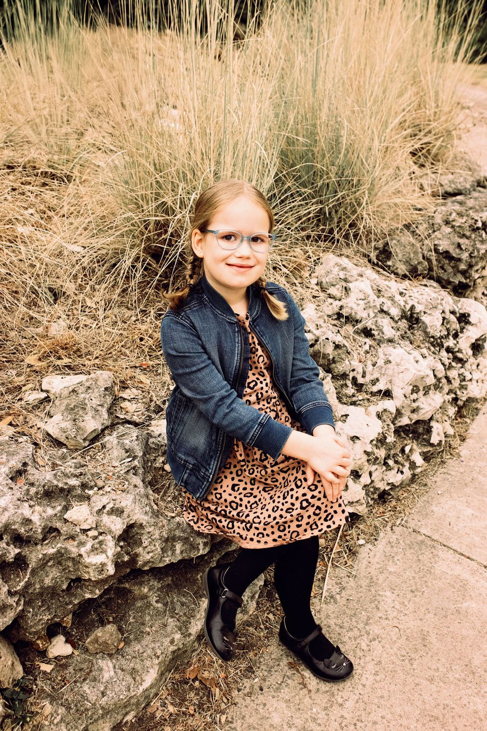 A young person in glasses sits on a rocky ledge wearing a denim jacket and leopard print dress against tall ornamental grasses.