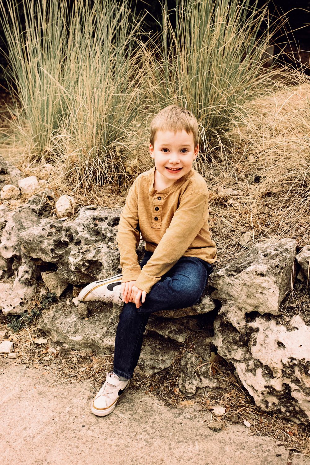 A toddler in a tan sweater and jeans sits casually on a stone wall with dry grass in the background.