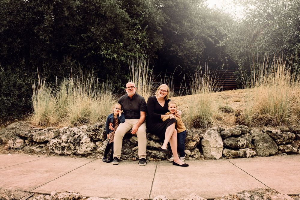 Two people sit on a stone wall along a path with tall ornamental grasses and trees in the background.