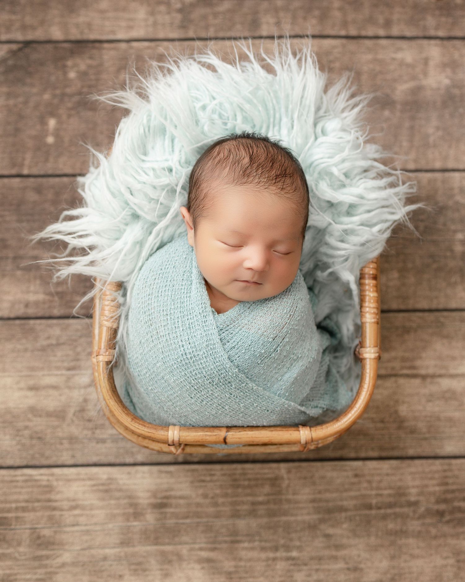 Sleeping newborn wrapped in soft blue fabric rests peacefully in wicker basket with fluffy white fur backdrop.