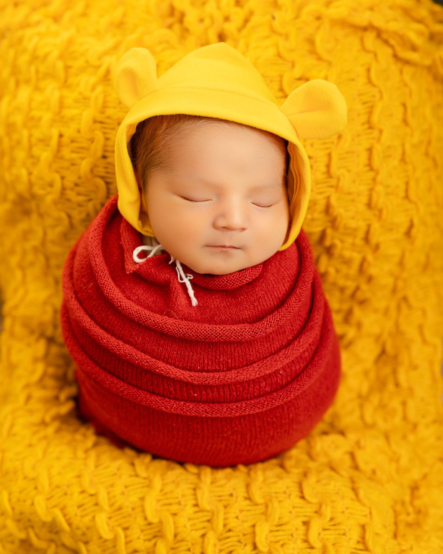 A sleeping baby wrapped in a red blanket wearing a yellow beanie hat against a bright yellow textured background.