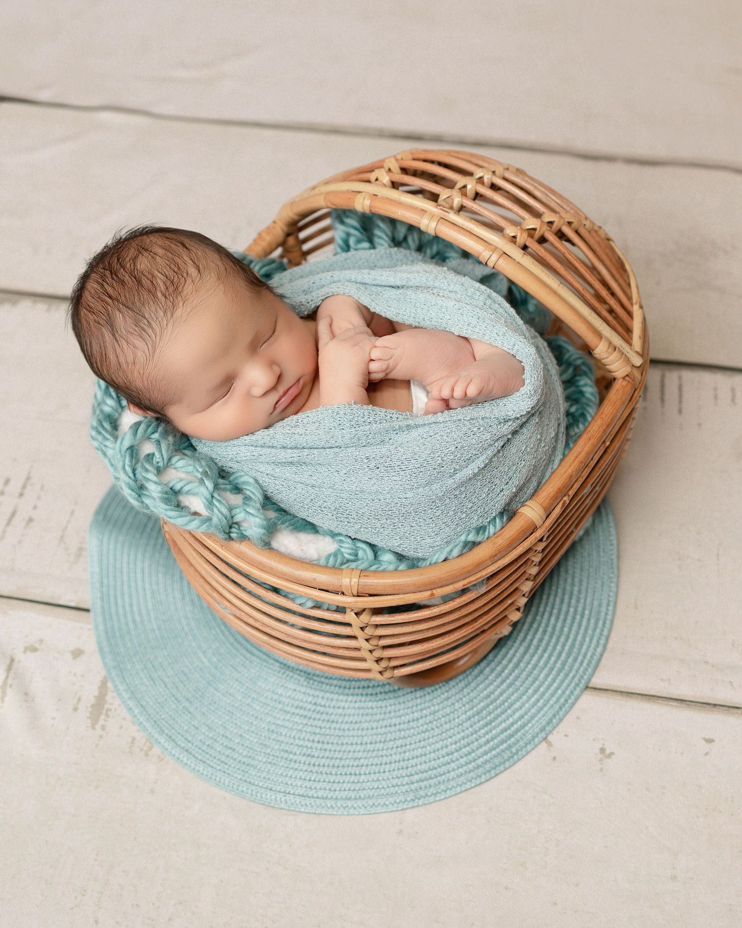 A sleeping newborn wrapped in soft turquoise fabric lies peacefully in a woven basket on a blue circular mat.