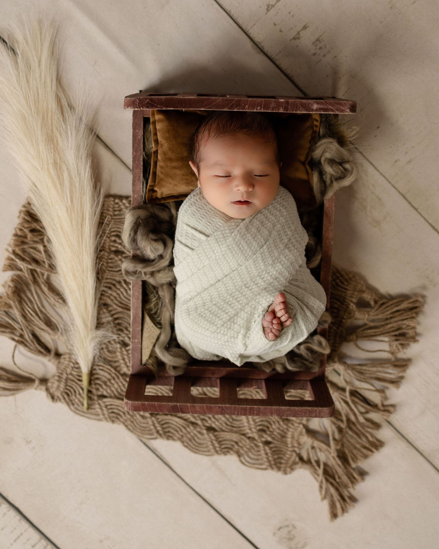 A sleeping infant swaddled in white lies in a rustic wooden box with pampas grass decor on a neutral floor.
