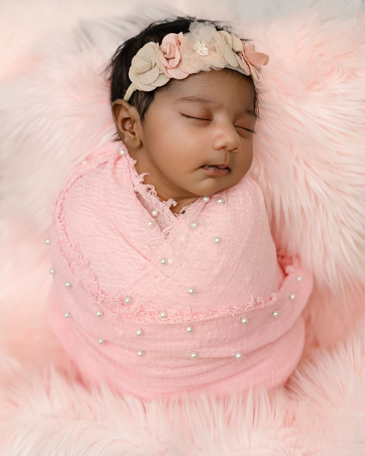 Peaceful sleeping newborn swaddled in pink wrap with floral headband on soft pink furry backdrop.