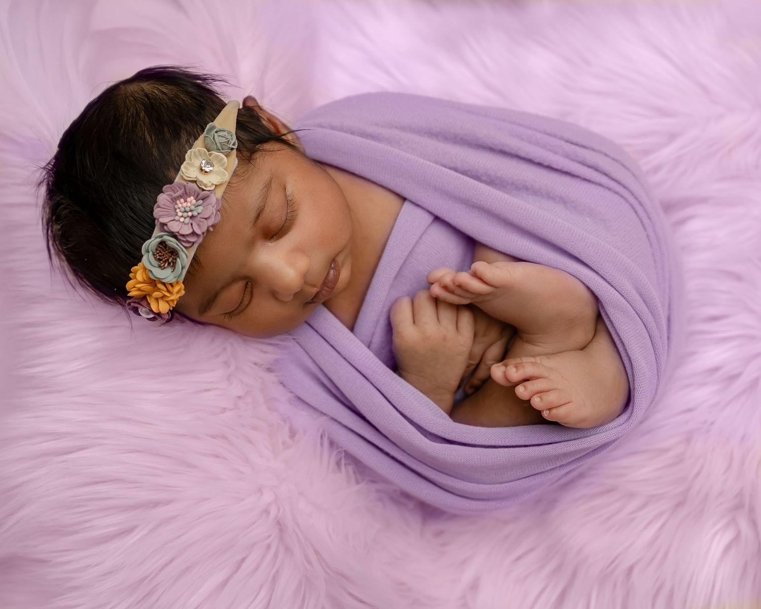 Sleeping newborn wrapped in soft purple fabric rests on fluffy pink blanket wearing delicate beaded headband.