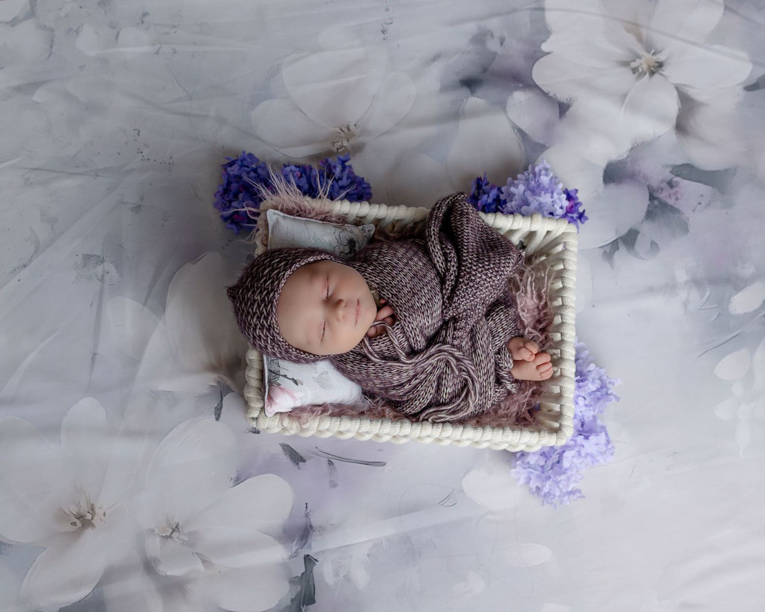 A sleeping baby wearing a gray knit sweater lies in a white basket with purple accents surrounded by soft textured fabric.