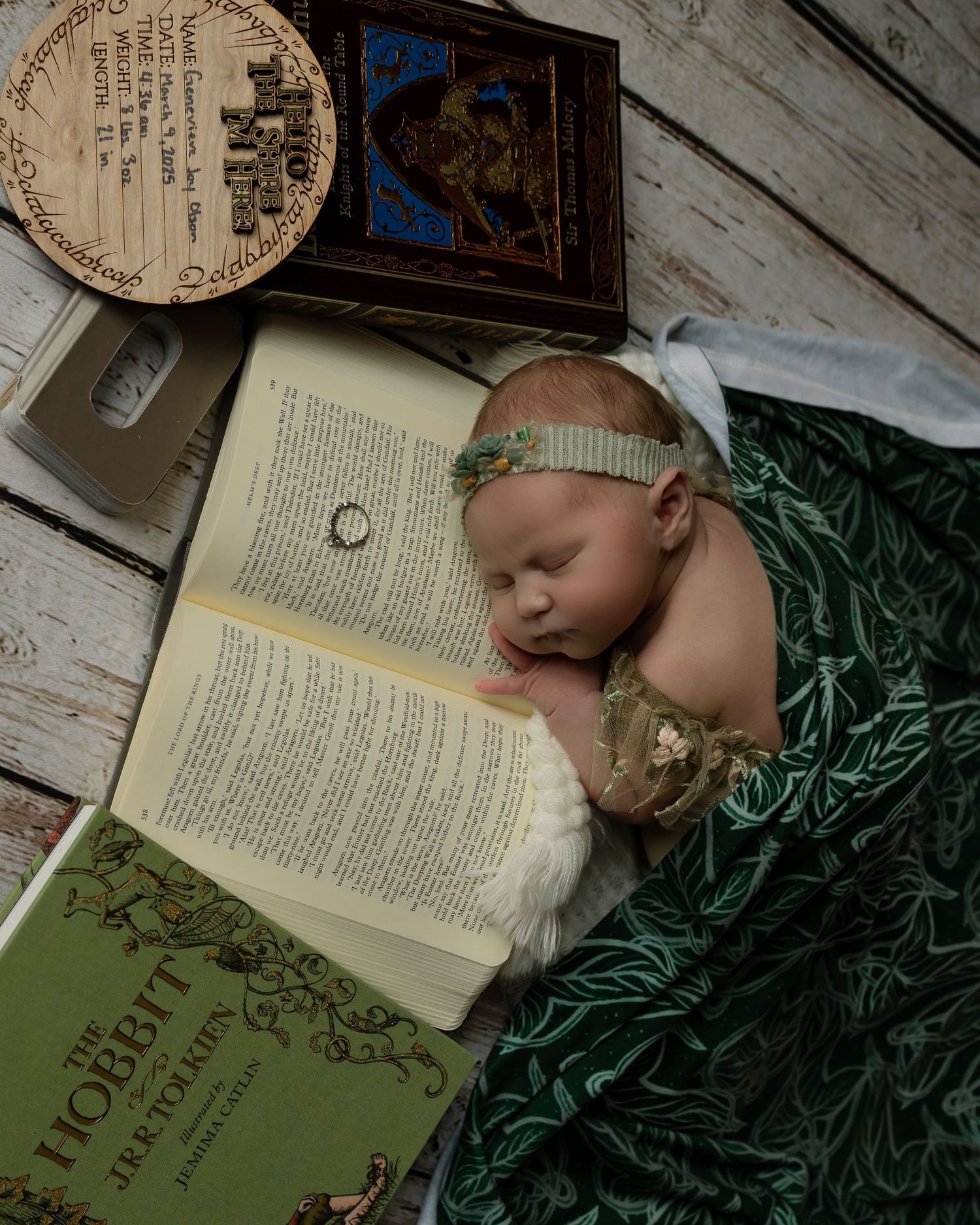 A sleeping baby wrapped in green fabric lies next to an open vintage book on white wooden floor.