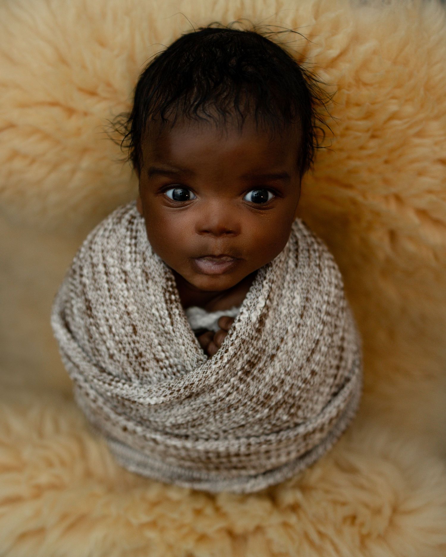 A baby wrapped in a cozy knitted ivory cowl scarf sits on a fluffy beige fur blanket.