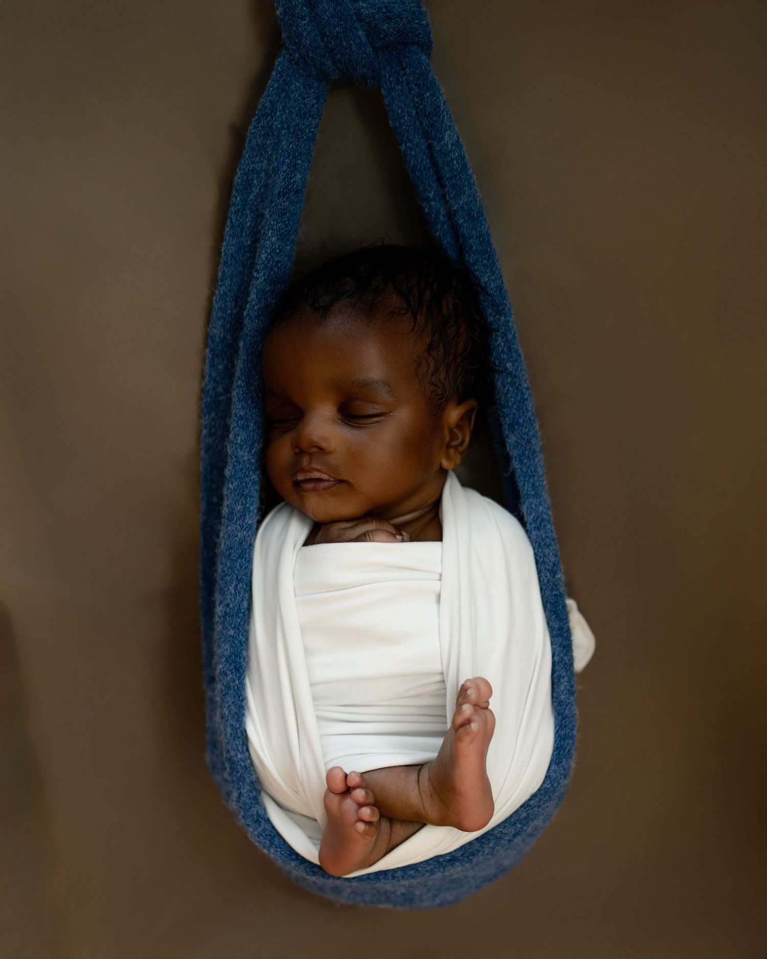 Sleeping infant rests peacefully in a blue fabric sling suspended against a neutral brown backdrop.