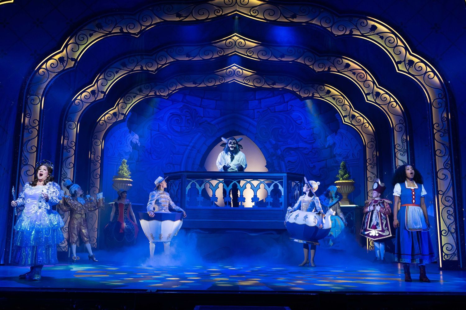 Ethereal stage scene with performers in white costumes under blue-lit Gothic arches creating mystical atmosphere.