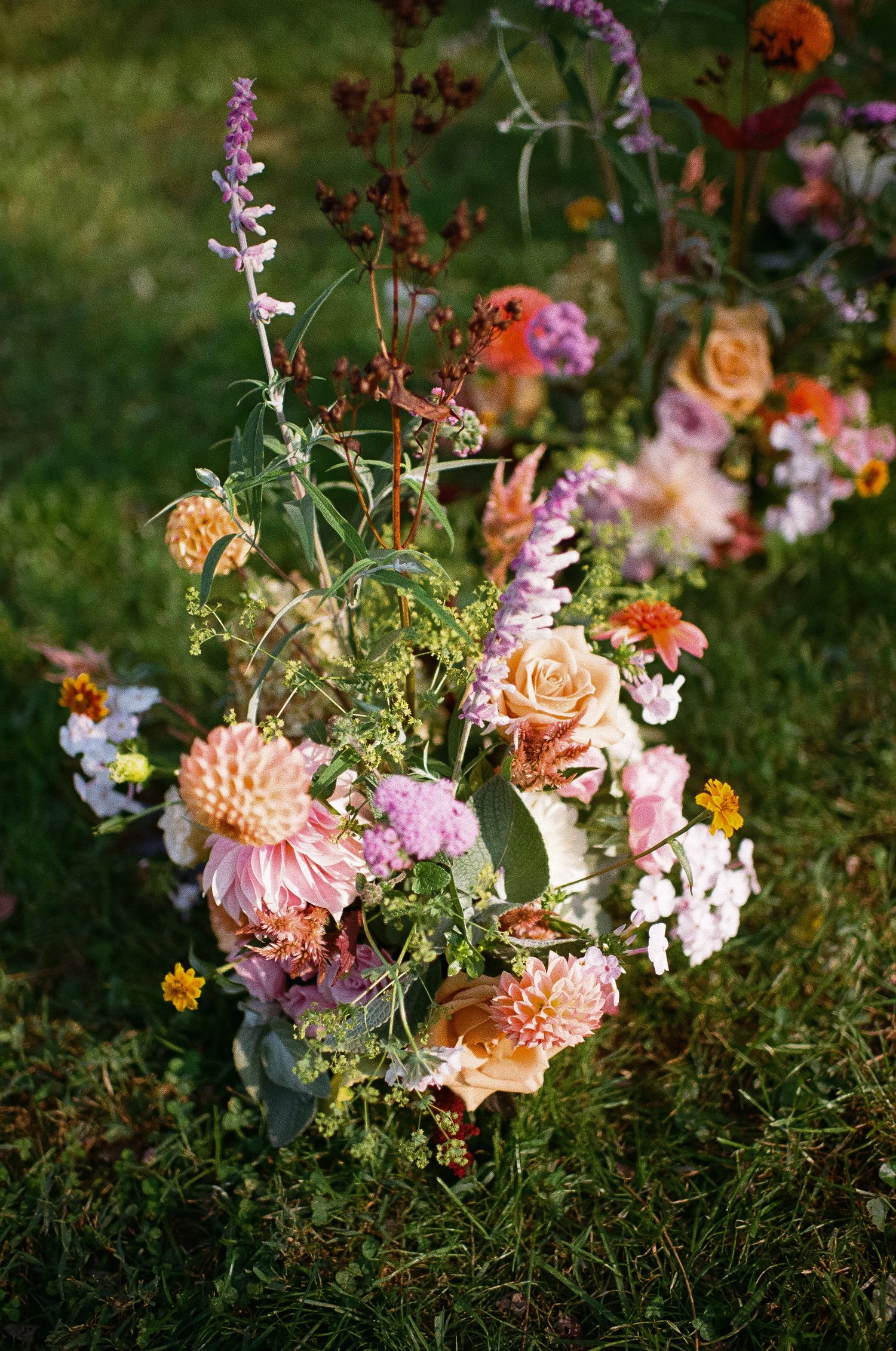 A romantic garden arrangement featuring peach roses, pink dahlias, and wildflowers set against green grass in soft light.