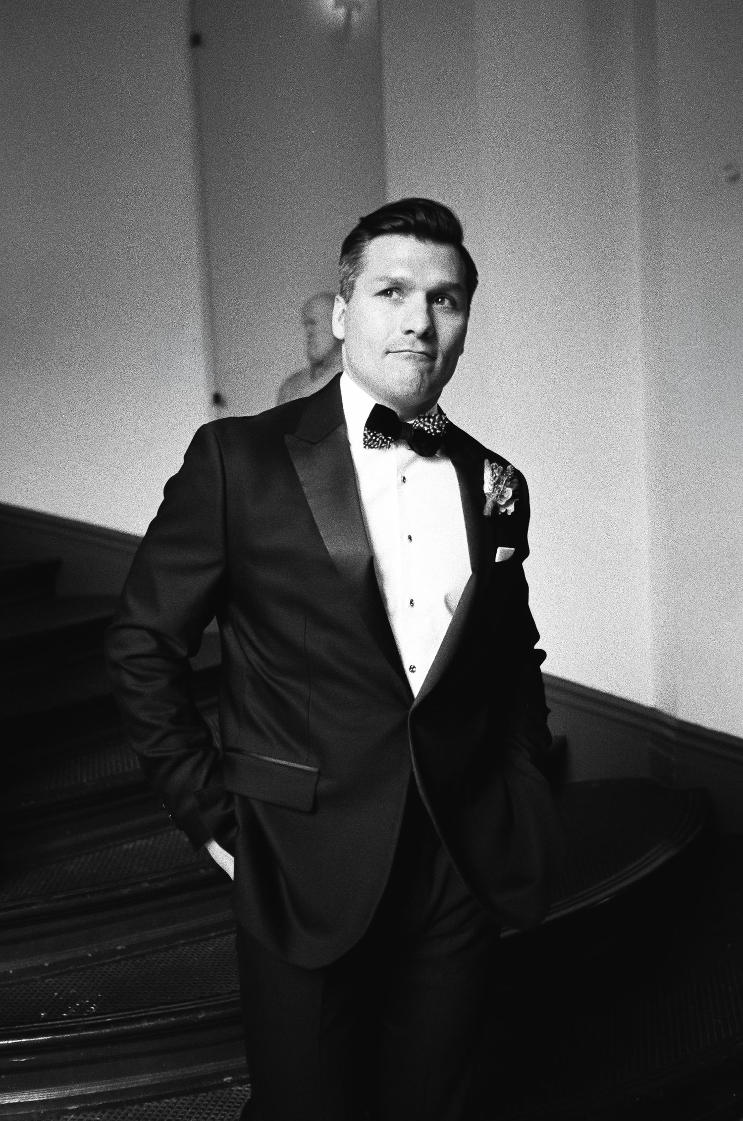 Black and white photo of a person in a formal tuxedo with bowtie and boutonniere standing beside a piano.