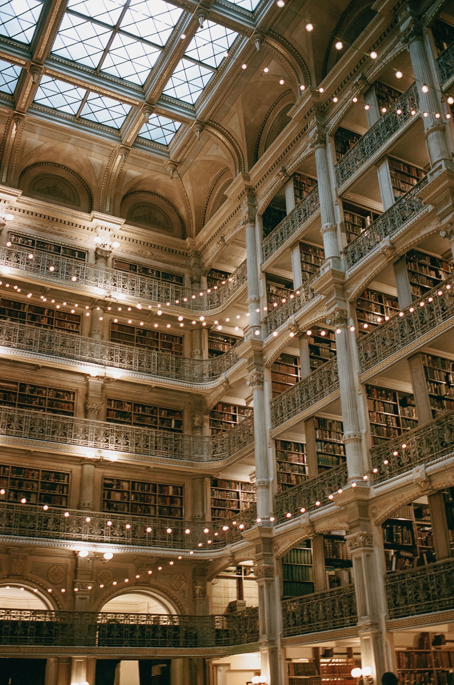 Grand interior of historic Peabody Library with ornate cast-iron balconies, glass ceiling, and multiple levels of book-lined shelves.