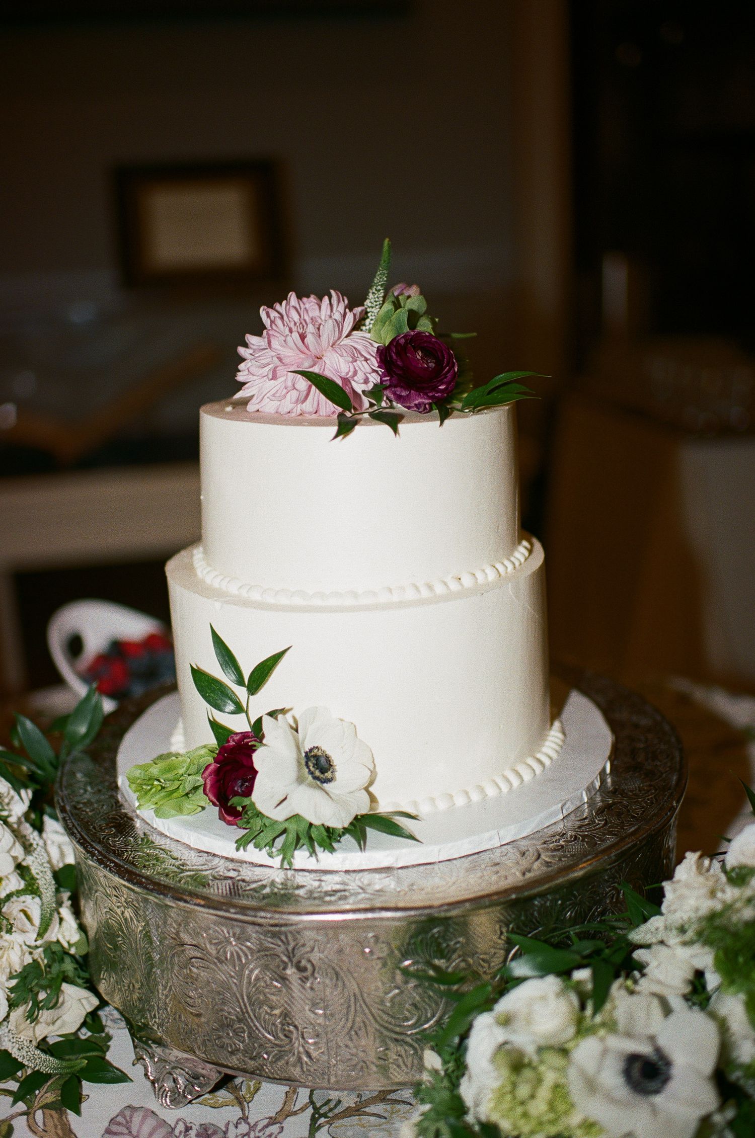 Elegant two-tier white wedding cake decorated with pink and burgundy flowers on ornate silver cake stand.