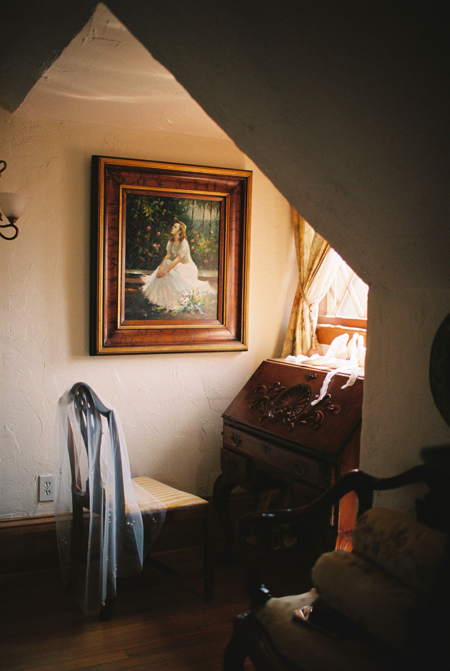 Vintage room corner with framed painting, blue fabric draped on chair, and wooden desk beneath slanted ceiling.