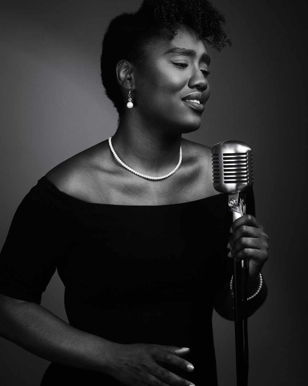 Elegant singer in off-shoulder black dress holds vintage mic, lit with moody side light and cinematic shadow detail.