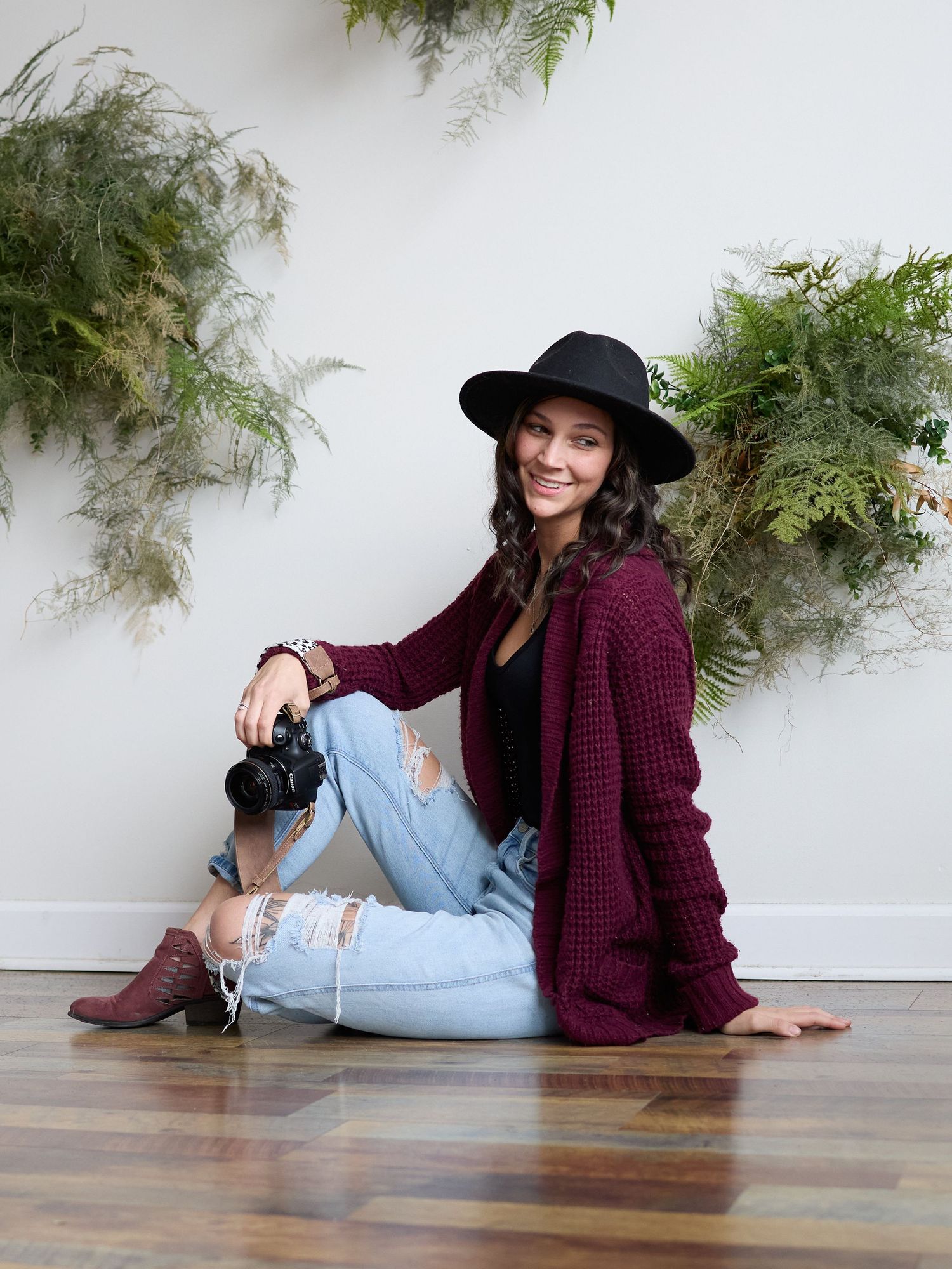 Person in red oversized cardigan and white sneakers sits on wooden stool holding camera for portrait photoshoot.