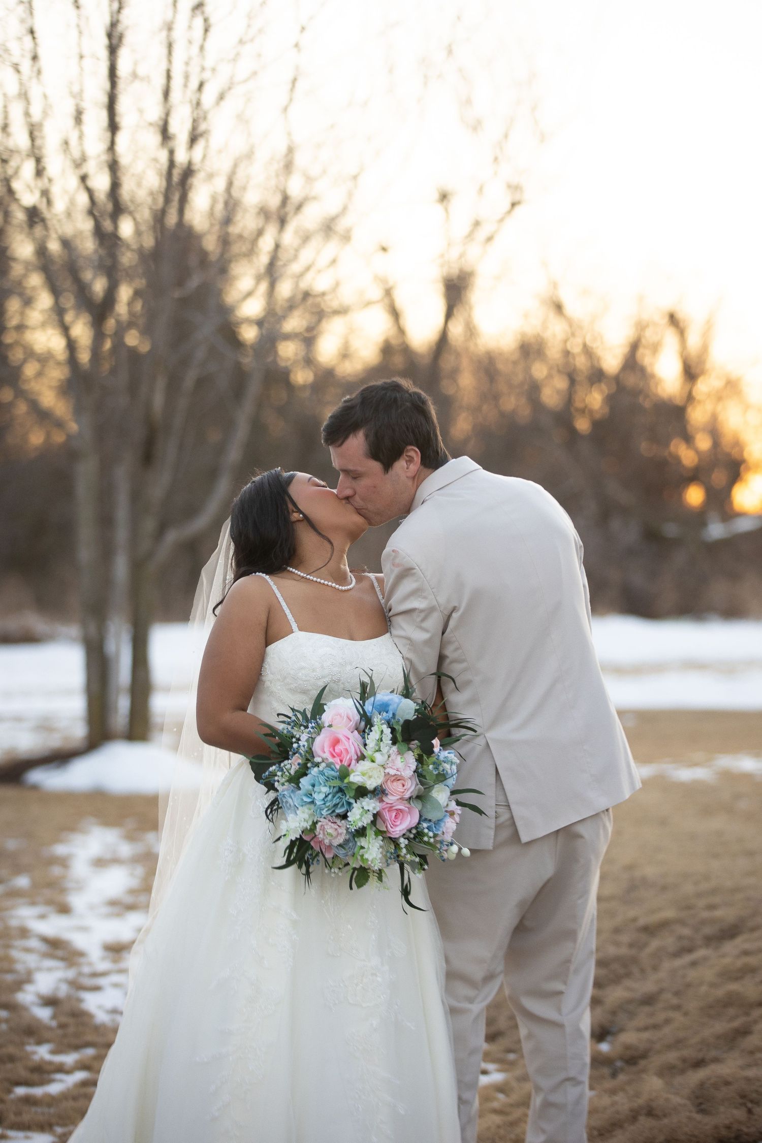 A winter wedding photo showing a couple embracing with a pastel bouquet at sunset.