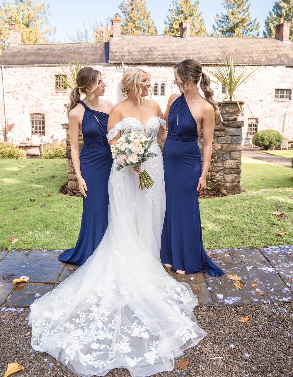 Bridal party in navy blue dresses and white gown posing outdoors near stone building.