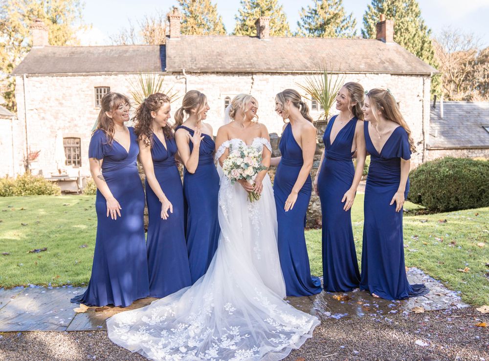 Bridesmaids in royal blue dresses pose with bride wearing lace gown in autumn garden setting.
