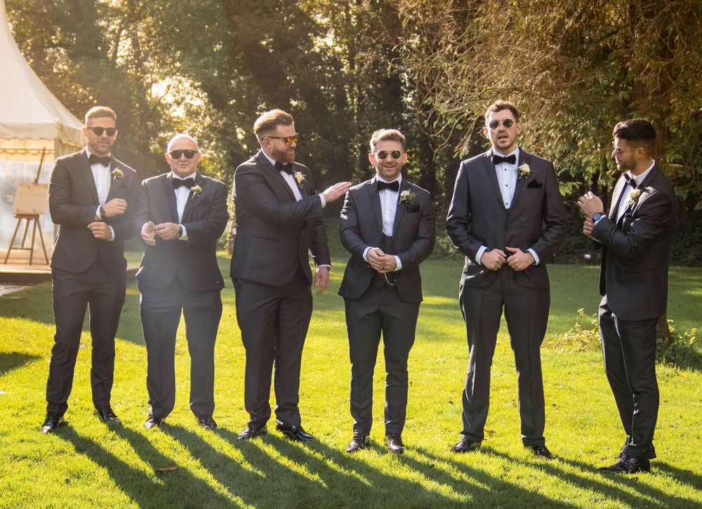 Wedding party groomsmen lined up outdoors in black tuxedos at sunset.