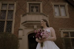 A bride holding a pink bouquet poses in front of a historic mansion.
