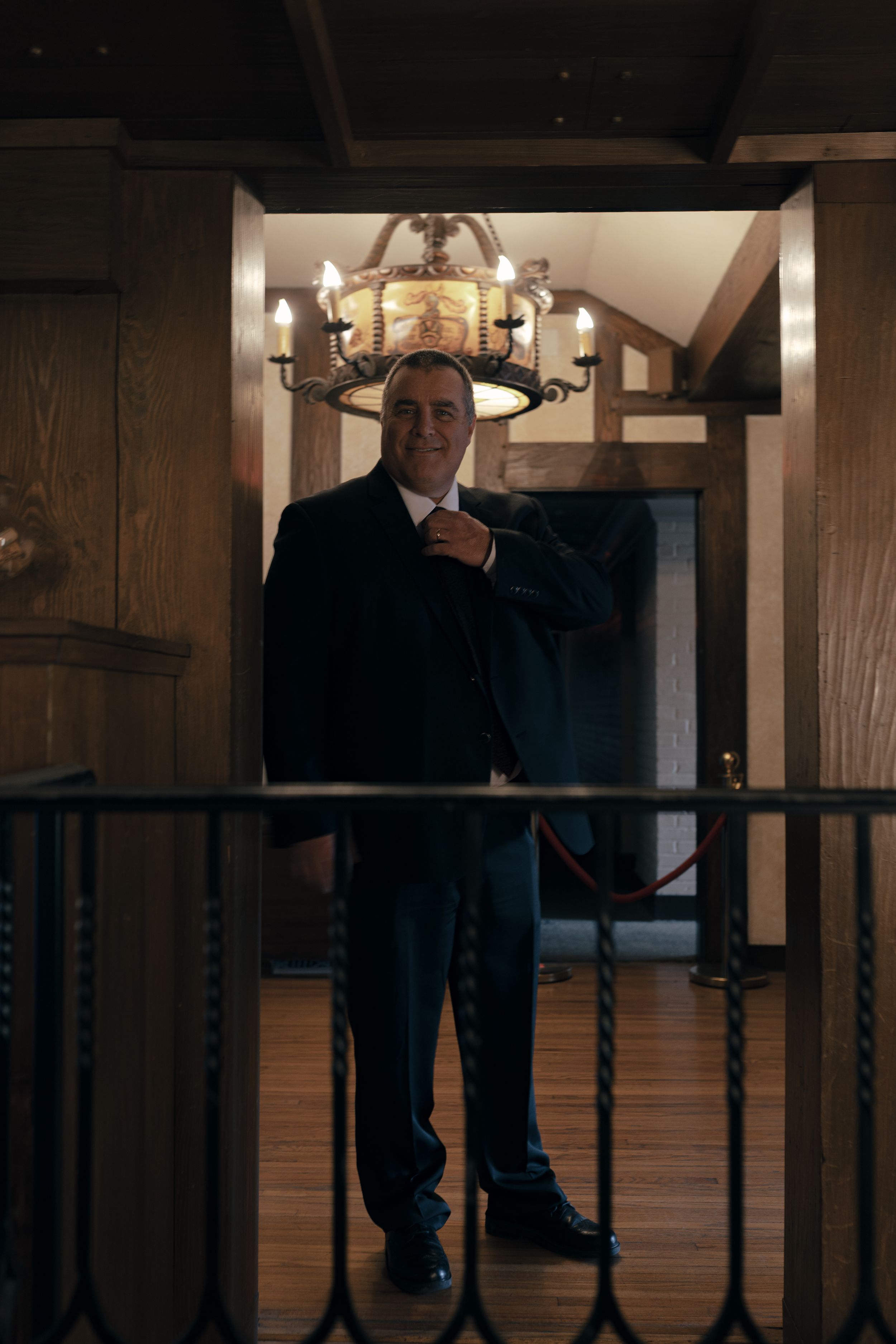 A formal portrait of someone standing on a wooden staircase beneath a chandelier.