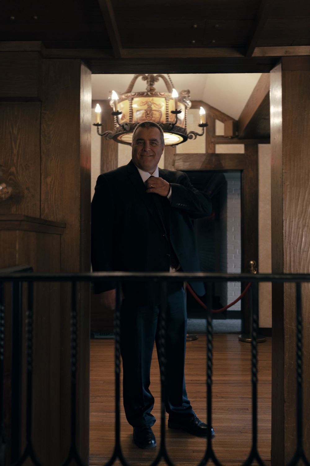 A formal portrait of someone standing on a wooden staircase beneath a chandelier.