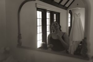 A wedding dress hangs in a window while a reflection appears in an arched mirror.