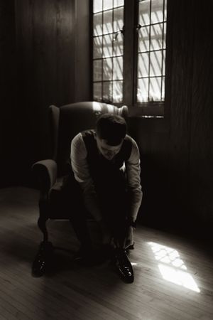 A person in formal attire sits alone in a dark room illuminated by window light.