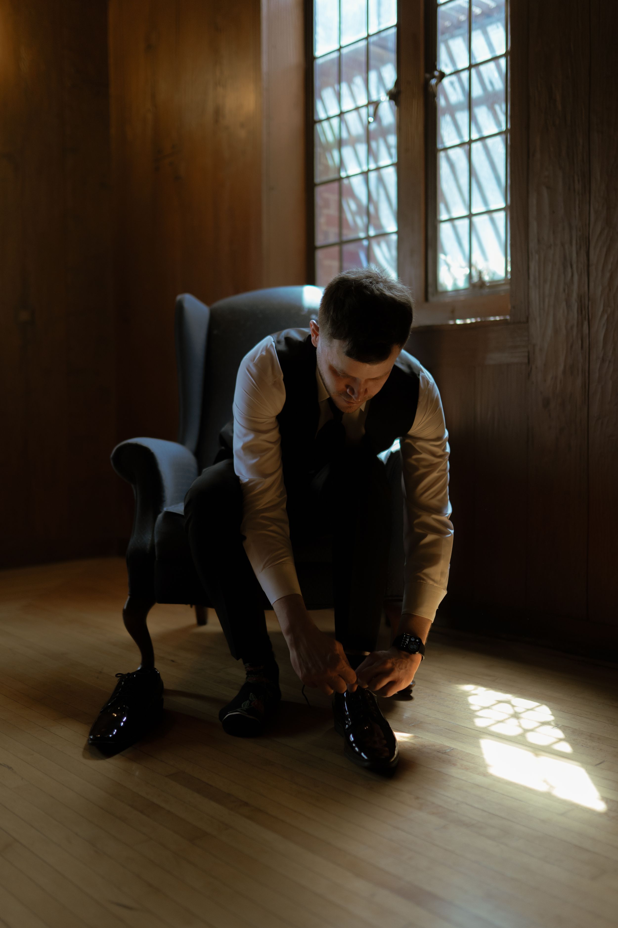 Figure in dark suit crouches down on wooden floor in sunlit room.