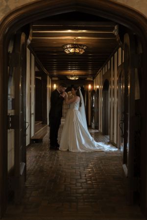 A romantic wedding photo in a dimly lit hallway with elegant chandeliers overhead.