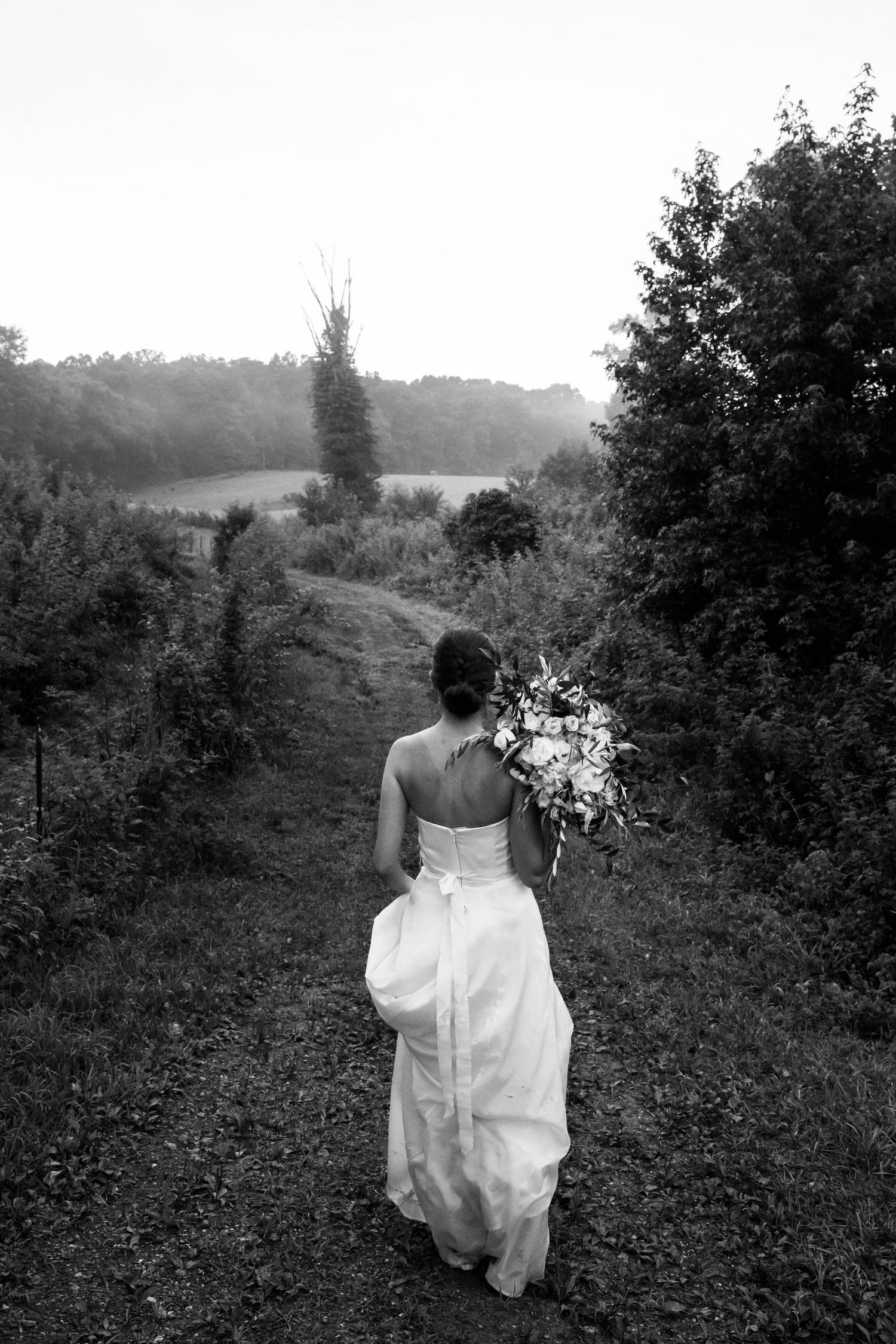 Black and white photo of a bride walking down a trail with her wedding bouquet thrown over her shoulder. Southwest Virginia photographer