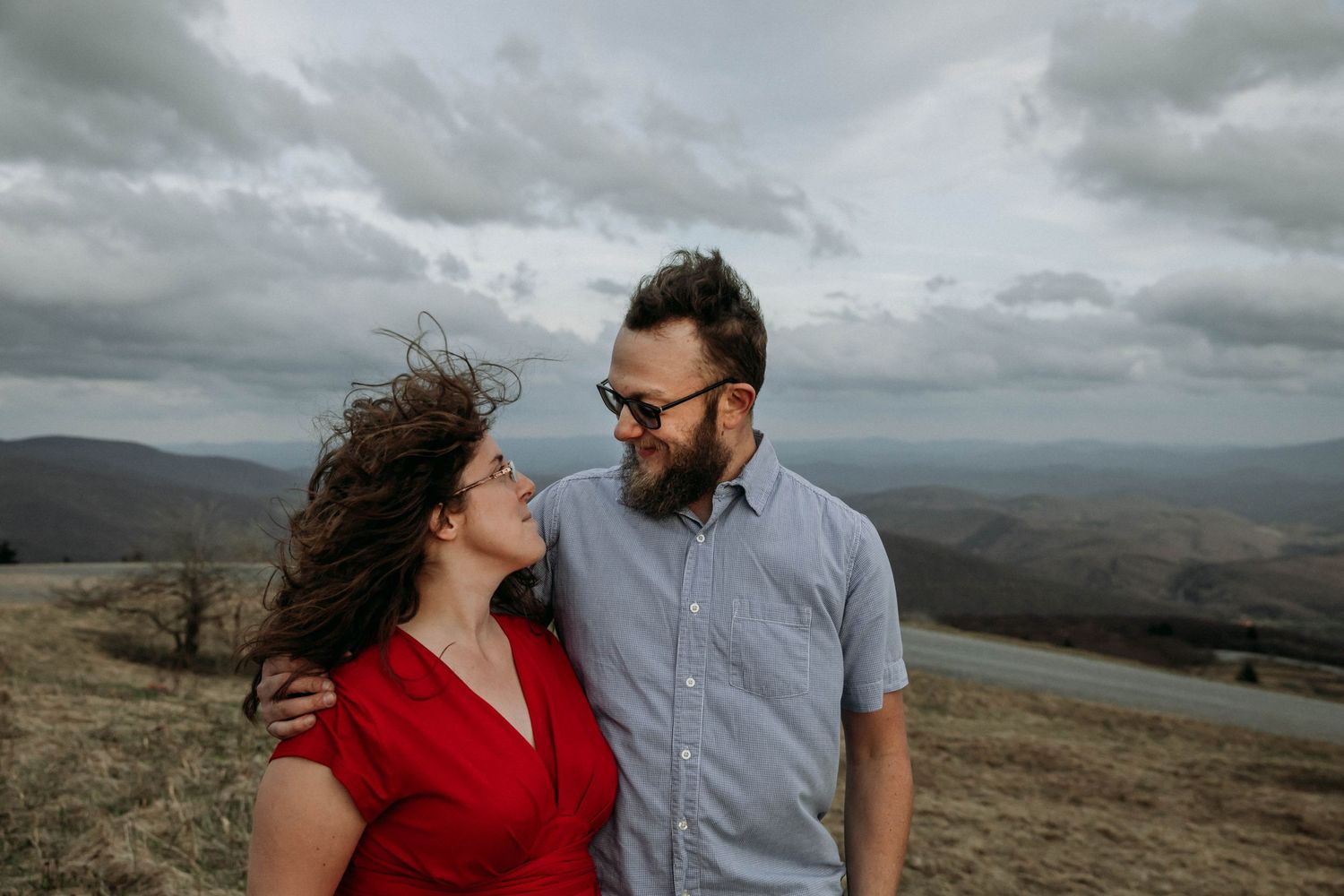 Photo of a man in a blue shirt with his arm around a woman in a red dress, looking at each other and smiling on Whitetop Mountain, Virginia