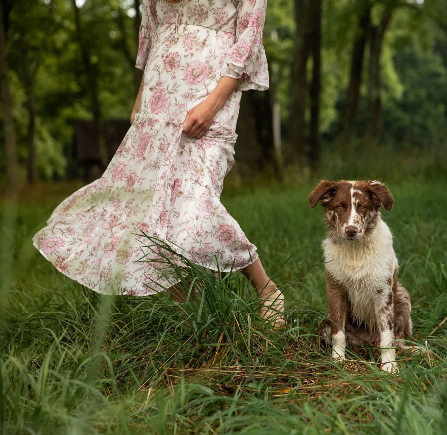 Photo of a woman in a floral dress, spinning around in a forest with a dog beside her.