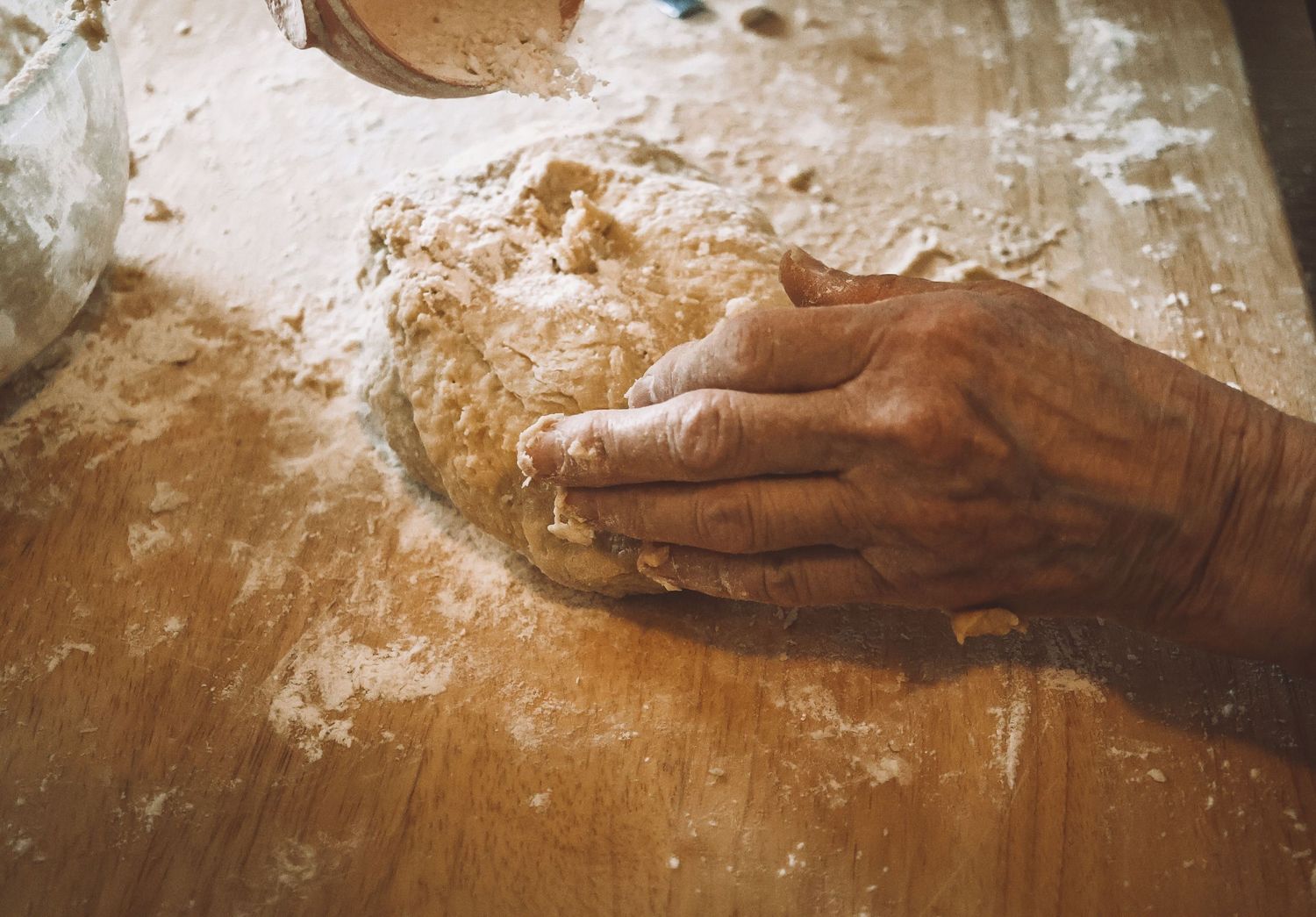 A hand dusted with flour kneads dough on a wooden surface while baking.