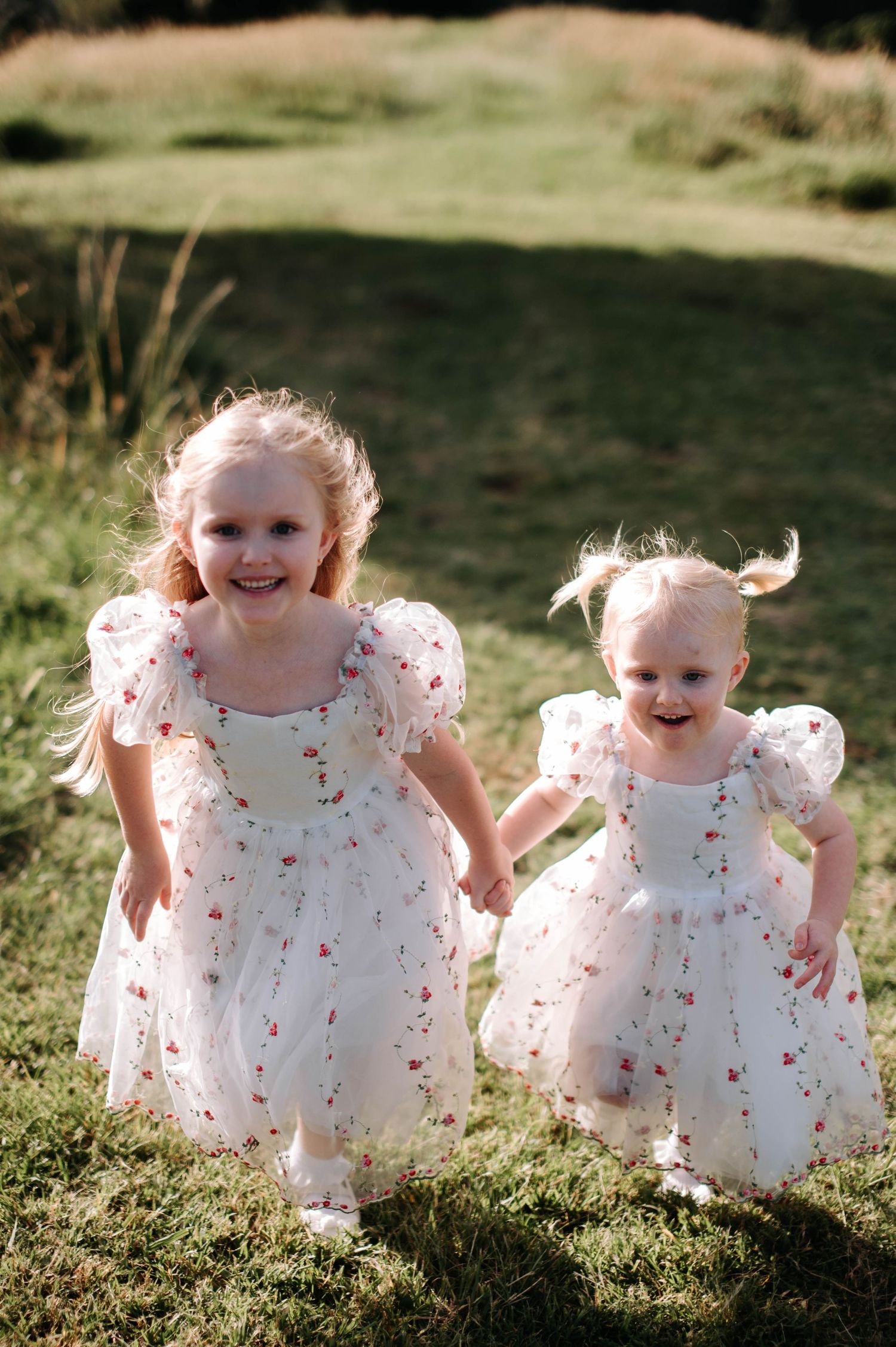 Two little girls in matching white floral dresses smiling in a sunny garden.
