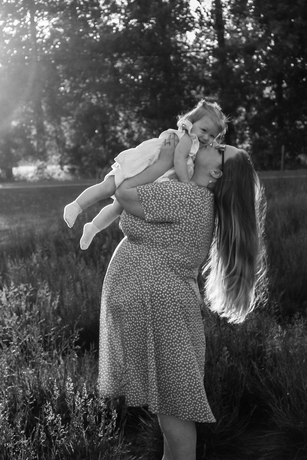 Black and white image of a mother lifting her laughing toddler in a sunlit outdoor field.